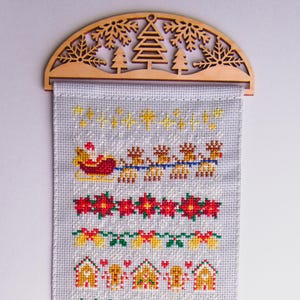 Pode incluir: Um bordado de ponto cruz branco com um tema natalino. O bordado apresenta um trenó vermelho e amarelo puxado por renas, uma fileira de poinsétias vermelhas, uma fileira de sinos amarelos e uma fileira de casas de gengibre.