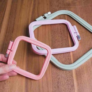Può includere: Una collezione di telai da ricamo quadrati nei toni del rosa e del verde. Un telaio rosa è tenuto in mano. I telai hanno un design liscio e arrotondato con una chiusura in metallo. Lo sfondo è una superficie in legno.
