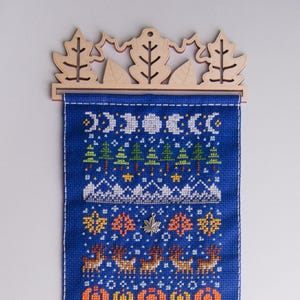 May include: A blue cross-stitch wall hanging with a winter forest scene. The hanging features a white snowy mountain range, a row of green trees, and a row of orange and yellow leaves. The hanging is attached to a wooden frame with a leaf design.