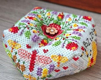 May include: A square, light blue fabric pincushion featuring intricate cross-stitch embroidery of colorful flowers, green vines, and dragonflies. A small, five-petaled wooden button is centered on top, and the letters BZ are stitched on the side.