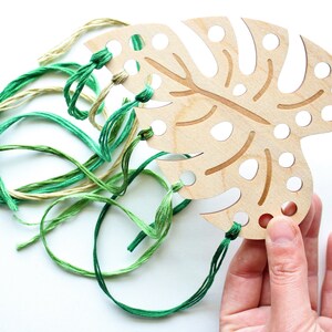 May include: A wooden monstera leaf with holes for stringing yarn. The leaf is light brown with a darker brown vein pattern. Green, yellow, and cream colored yarn is threaded through the holes.