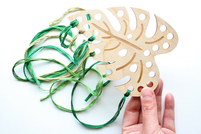 May include: A wooden monstera leaf with holes for stringing yarn. The leaf is light brown with a darker brown vein pattern. Green, yellow, and cream colored yarn is threaded through the holes.