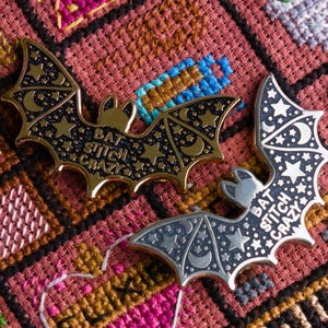 May include: Two gold and silver bat-shaped pins with the text "Bat Stitch Crazy" on a red and brown fabric background.