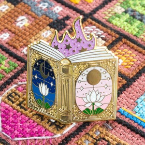 以下が含まれることがあります： 金色のエナメルピン。開いた本を模しており、左ページは月と星の夜のシーン、右ページは太陽と星を描いています。紫色の炎と星が本の上にあります。クロスステッチの背景に配置されています。