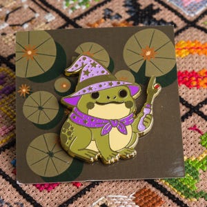 May include: A whimsical enamel pin featuring a green frog wizard with a purple hat and scarf, holding a wand. The pin is set on a brown card with lily pad designs. The frog has gold accents.
