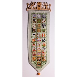 May include: A Halloween cross-stitch banner with a wooden topper. The banner displays embroidered squares with spooky images, including bats, cats, and pumpkins. The year "2025" is stitched at the bottom, and an orange bead hangs from the point.