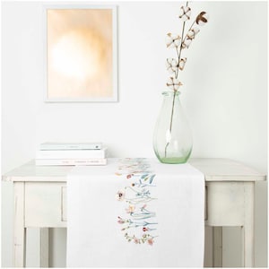 May include: A white table runner with embroidered floral designs in pastel colours. A clear glass vase holds a cotton stem. A framed artwork and stacked books are also on the white table.