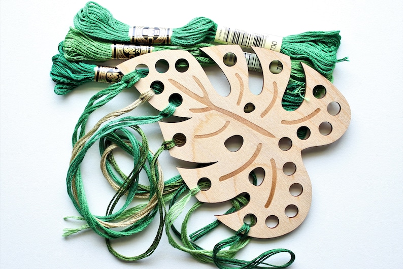 May include: A wooden monstera leaf shaped embroidery hoop with green and beige embroidery floss threaded through the holes.