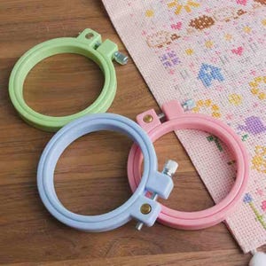 Puede incluir: Tres bastidores de bordado de plástico en colores pastel: verde claro, azul claro y rosa. Cada bastidor tiene un tornillo plateado para apretar. Un trozo de tela de punto de cruz con un patrón floral es parcialmente visible.