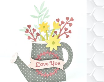 Puede incluir: Una regadera gris con lunares blancos y una cinta roja y blanca que dice "Love You". La regadera está llena de un ramo de flores amarillas y hojas verdes.