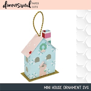 May include: A blue and pink paper house ornament with a gold glitter base and a red and white striped chimney. The house has a wreath on the front and a star on the side. "MINI HOUSE ORNAMENT SVG" is written at the bottom of the image.