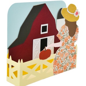 Puede incluir: Un granero rojo con una puerta blanca y una valla amarilla. Una mujer con un vestido floral y un sombrero de paja sostiene una calabaza frente al granero.