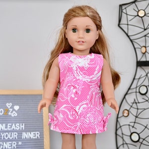 May include: A pink and white patterned dress with a butterfly design for a doll. The dress has a sleeveless design and a bow on each side. The doll has long blonde hair and blue eyes.