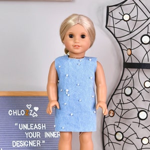 May include: A doll wearing a light blue denim dress with white pearl accents. The doll has blonde hair and blue eyes. The doll is standing on a white surface in front of a gray letter board with the text "CHLOZZ "UNLEASH YOUR INNER DESIGNER"".