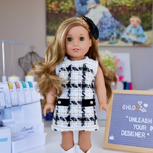 May include: A doll wearing a white and black checkered dress with gold accents and white cowboy boots. The doll has long brown hair and blue eyes. The doll is standing on a white surface in front of a sewing machine and a sign that says "Chlozz "UNLEASH YOUR INNER DESIGNER"