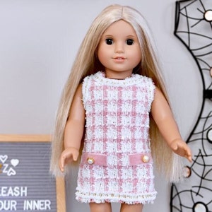 May include: A doll with long blonde hair wearing a sleeveless pink and white plaid dress. The dress has a pink velvet belt with pearl accents and gold trim. The doll is standing in front of a gray felt letter board with the text "UNLEASH YOUR INNER".