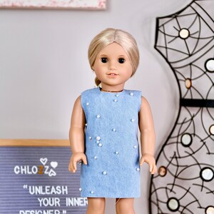 May include: A doll wearing a light blue denim dress with white pearl embellishments. The doll has blonde hair and blue eyes. The doll is standing on a white surface in front of a black metal frame and a sign that reads "CHLOZZ "UNLEASH YOUR INNER DESIGNER""