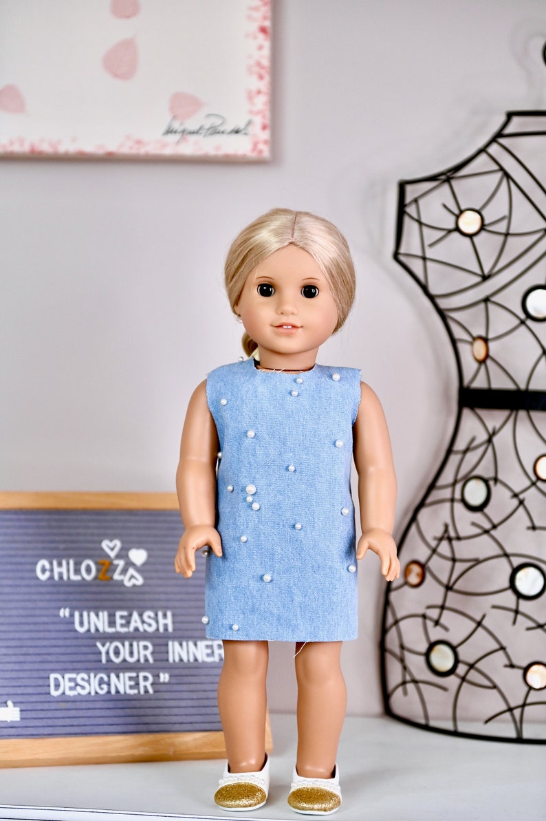 May include: A doll wearing a light blue denim dress with white pearl embellishments. The doll has blonde hair and blue eyes. The doll is standing on a white surface in front of a black metal frame and a sign that reads "CHLOZZ "UNLEASH YOUR INNER DESIGNER""
