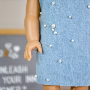 May include: A light blue denim dress with white pearl embellishments. The dress is sleeveless and has a simple, straight silhouette.