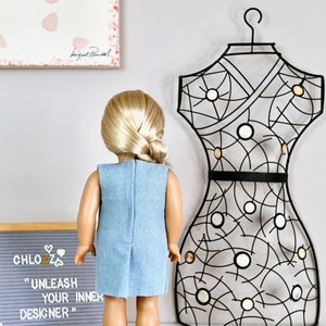 May include: A doll wearing a light blue denim dress with a black belt. The doll has blonde hair in a ponytail and is standing in front of a black metal dress form. A sign behind the doll reads "CHLOZZ "UNLEASH YOUR INNER DESIGNER""