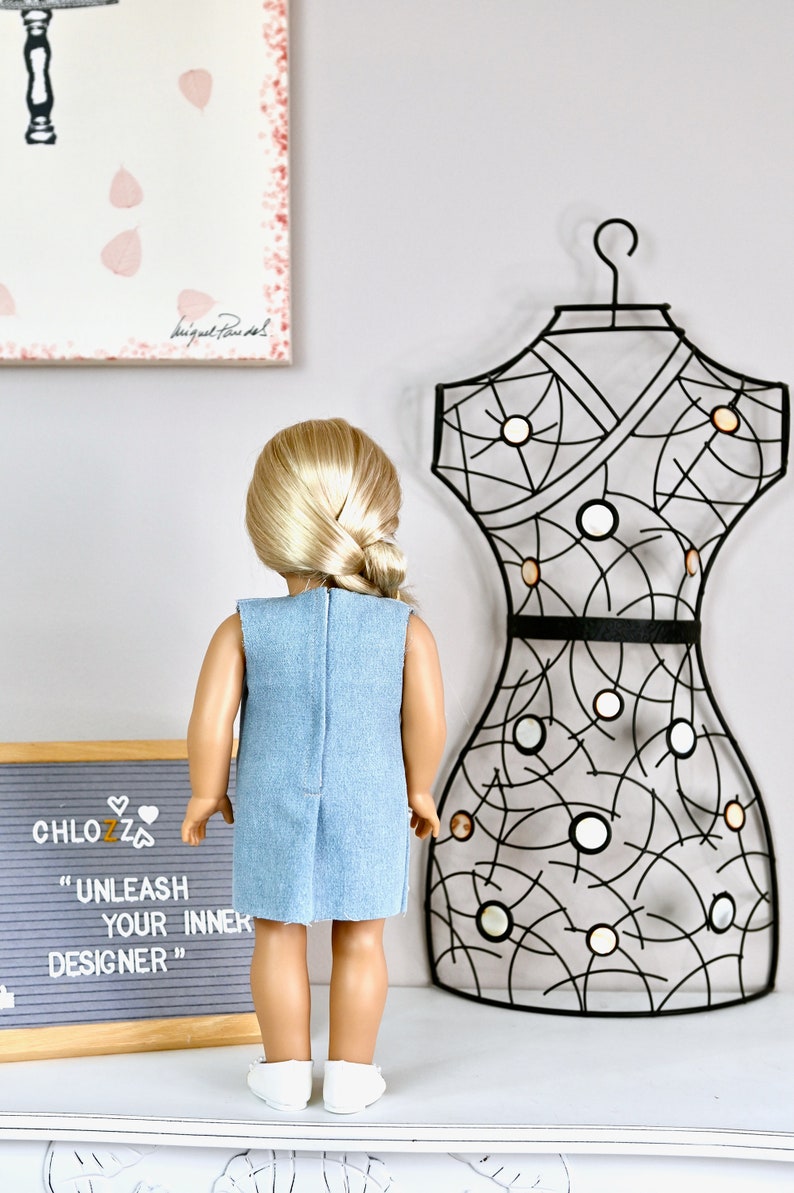 May include: A doll wearing a light blue denim dress with a black belt. The doll has blonde hair in a ponytail and is standing in front of a black metal dress form. A sign behind the doll reads "CHLOZZ "UNLEASH YOUR INNER DESIGNER""