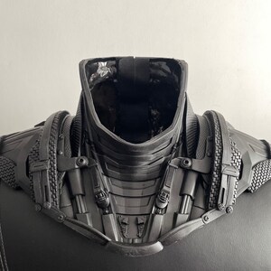 May include: Black and gray futuristic-style costume neck piece with a textured surface and multiple straps and buckles.