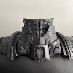 May include: Black plastic costume neck piece with a textured surface and a series of interlocking panels. The piece has a raised, textured design on the front and back.