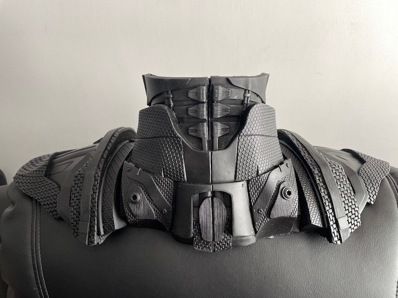 May include: Black plastic costume neck piece with a textured surface and a series of interlocking panels. The piece has a raised, textured design on the front and back.