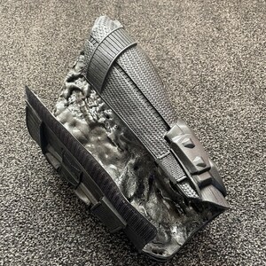 May include: Black plastic arm armor with a textured surface and a raised design. The armor is designed to look like a futuristic or fantasy piece of equipment.