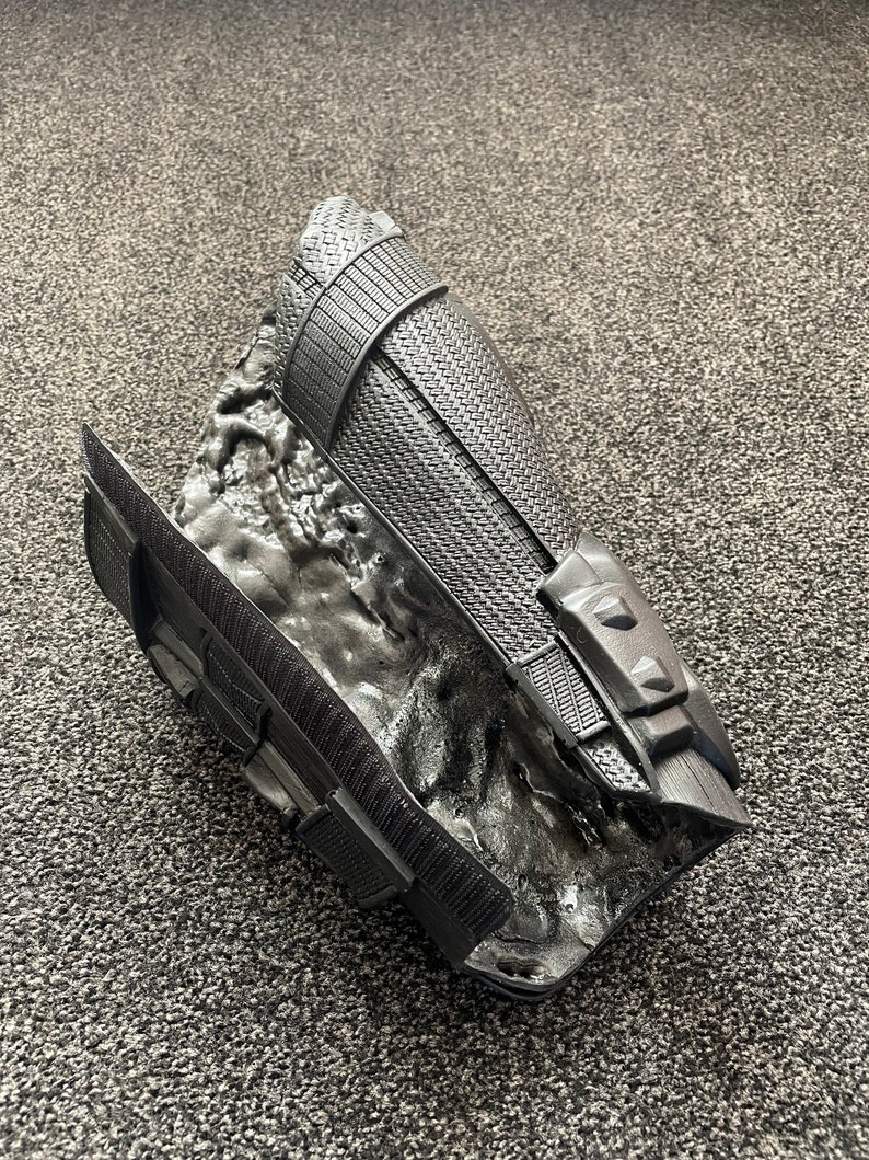 May include: Black plastic arm armor with a textured surface and a raised design. The armor is designed to look like a futuristic or fantasy piece of equipment.