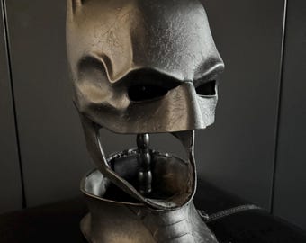 May include: A metallic gray Batman cowl, featuring pointed ears and a stylized facial design. The cowl is mounted on a stand, showcasing the full head and neck coverage. The overall design is reminiscent of the iconic superhero.
