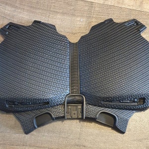 May include: Black plastic chest piece with a textured surface and a raised design. The chest piece has two small, round openings on the bottom.