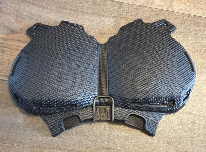May include: Black plastic chest piece with a textured surface and a raised design. The chest piece has two small, round openings on the bottom.