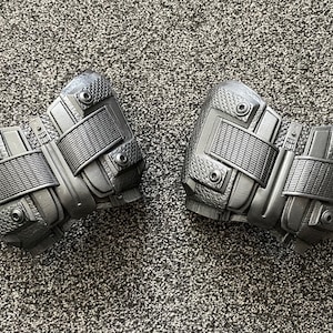 May include: A pair of black, textured, and detailed knee pads with straps and buckles.