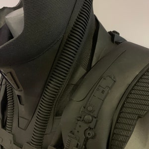 May include: A close-up of a black sculpture, possibly a bust or part of a costume. The piece has a textured, ribbed neck and shoulder area, with intricate mechanical details and a matte finish. The overall design suggests a futuristic or sci-fi theme.