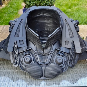 May include: A black 3D printed costume piece with a futuristic design. The piece has a large, open collar and intricate details, including a grid pattern and small circular elements.