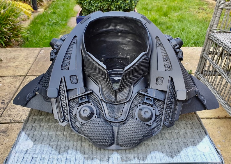 May include: A black 3D printed costume piece with a futuristic design. The piece has a large, open collar and intricate details, including a grid pattern and small circular elements.