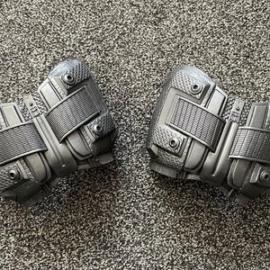May include: A pair of black, textured, and detailed knee pads with straps and buckles.