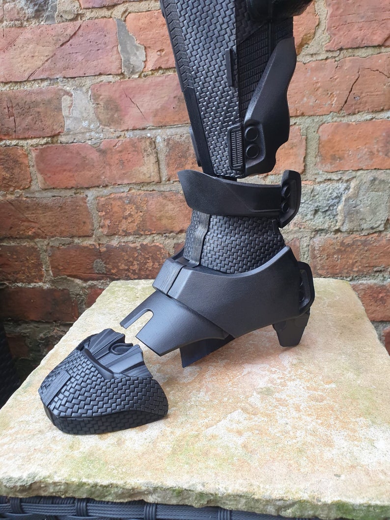 May include: Black plastic leg armor with a textured surface. The armor is in multiple pieces and is disassembled.
