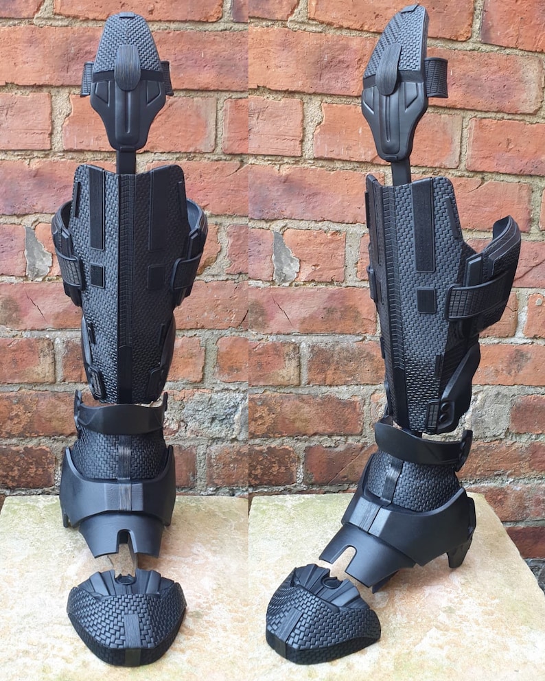 May include: A black, segmented, articulated leg armor piece, made of plastic, with a textured surface. The armor is in multiple pieces, and is shown in two different positions.