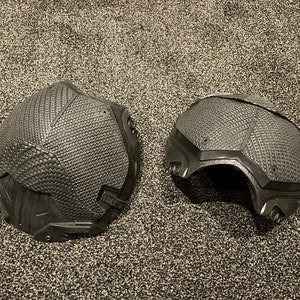 May include: Two black, textured, helmet-like pieces, possibly for cosplay or costume use. The pieces are made of a hard, molded material and have a geometric design.