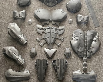 May include: A set of 3D printed armor pieces in a dark gray color. The pieces are laid out on a gray surface and include a helmet, chest plate, shoulder pads, arm guards, leg guards, and other smaller pieces.