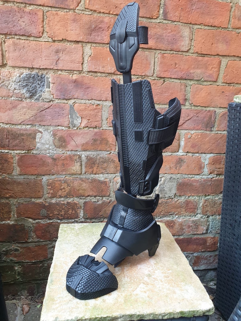 May include: Black plastic leg armor with a textured surface. The armor is in multiple pieces and is assembled together. The armor is on a light brown surface.