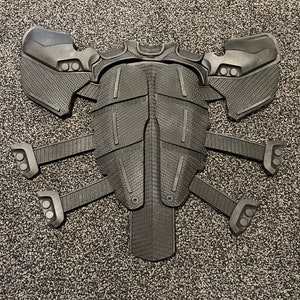 May include: A black plastic costume piece with a textured surface and multiple segmented panels. The piece has a symmetrical design with a central section and four extending arms.