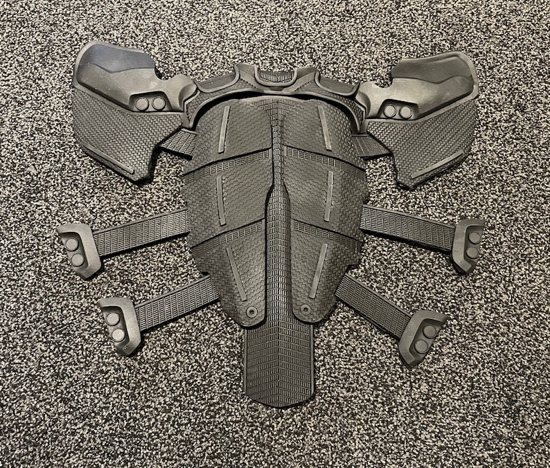 May include: A black plastic costume piece with a textured surface and multiple segmented panels. The piece has a symmetrical design with a central section and four extending arms.