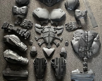 May include: A disassembled, dark gray, futuristic-looking armor set. The pieces include a helmet, chest plate, shoulder pads, gauntlets, leg armor, and other components, all with textured surfaces. The armor is arranged on a gray surface.