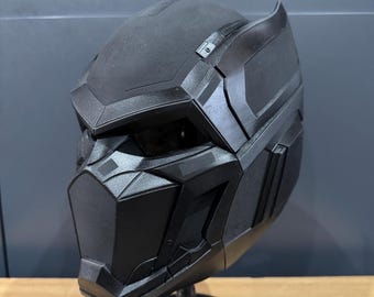 May include: A black, futuristic-style helmet with a textured surface. The helmet features angular designs, a face mask, and a raised section at the top. It is displayed on a black stand, showcasing its intricate details and design.