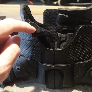May include: Black plastic armor with a honeycomb pattern and velcro straps. The armor is designed to be worn on the body and has a textured surface.