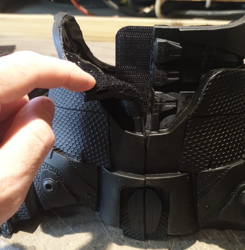 May include: Black plastic armor with a honeycomb pattern and velcro straps. The armor is designed to be worn on the body and has a textured surface.
