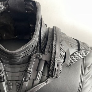 May include: Black and gray plastic costume piece with a textured surface and a series of buckles and straps. The piece appears to be a neck and shoulder armor piece.
